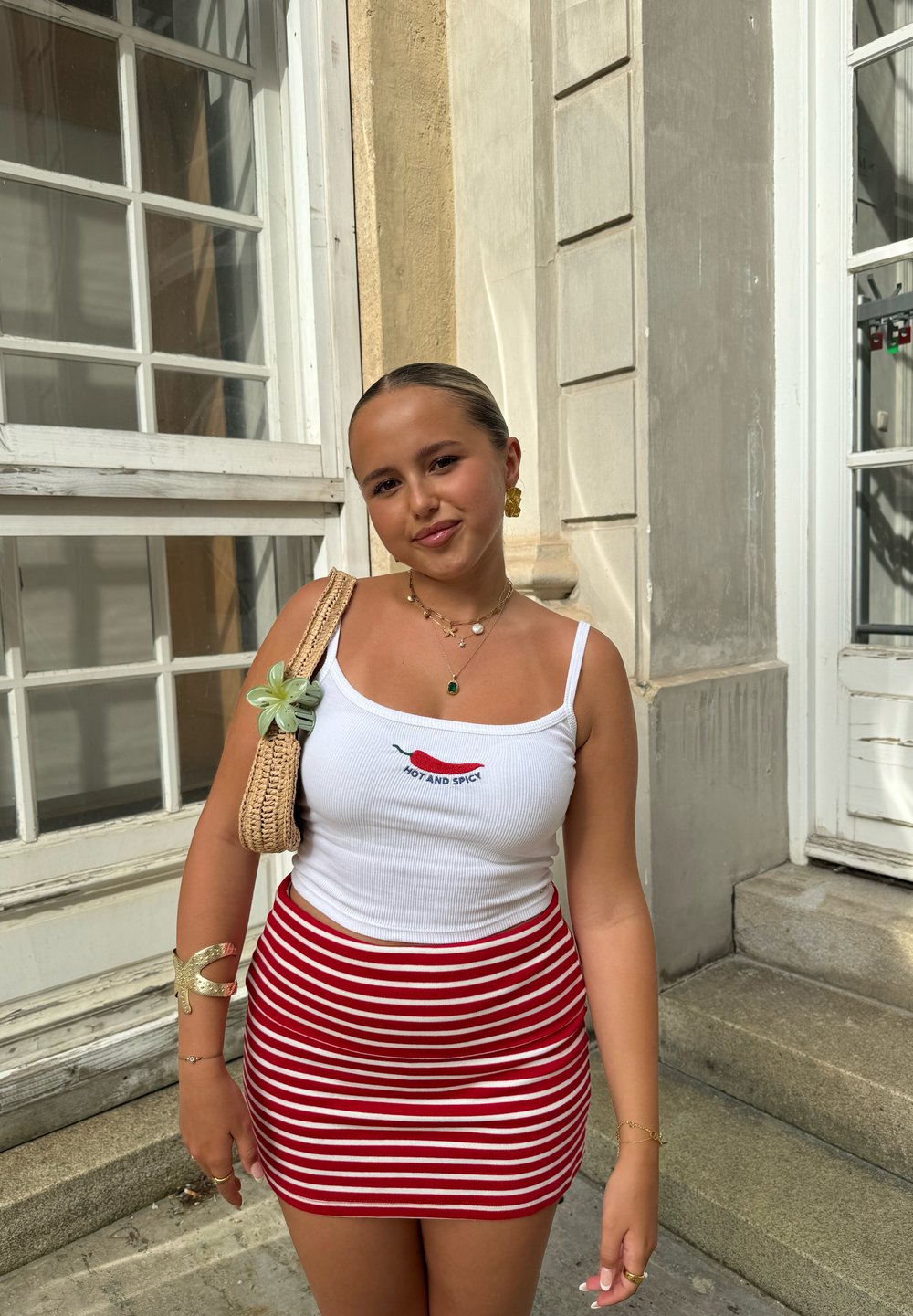 Top a maniche lunghe bianco a coste con la scritta "HOT AND SPICY" e grafica di peperoncino, abbinato a una mini gonna a righe rosse e bianche. Borsa intrecciata e accenti di gioielli.