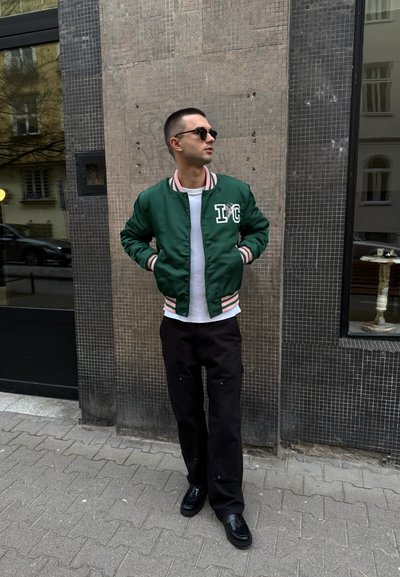 Chaqueta bomber de satén verde con puños y cuello a rayas, camiseta blanca, pantalones anchos negros y zapatos negros. Entorno urbano con azulejos texturizados.