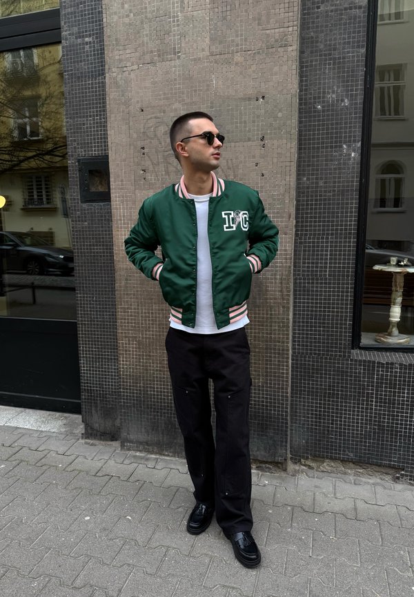 Groene satijnen bomberjack met gestreepte manchetten en kraag, witte T-shirt, zwarte wijde broek en zwarte schoenen. Stedelijke omgeving met gestructureerde tegels.
