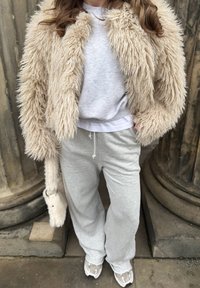 Personne portant une veste en fausse fourrure beige à poils longs, un pantalon de survêtement gris, des baskets gris clair, et tenant un sac à main assorti en fausse fourrure beige.