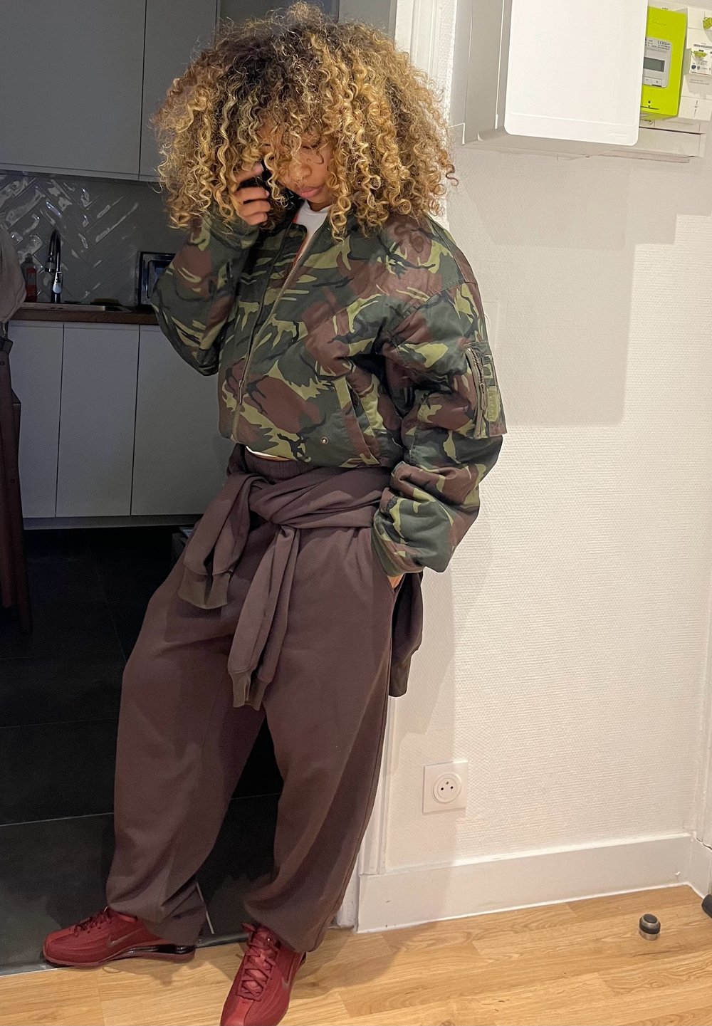 Casaco bomber camuflado em verde e castanho, combinado com calças largas castanhas e ténis vermelhos. Cabelo encaracolado, encostado numa parede.