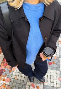 Must nööbitest jope, helesinine kampsun, tumedad laiad püksid ja kohvitassi hoidmine. Lehemustriga põrand. Vabaaja linnastiilne riietus.