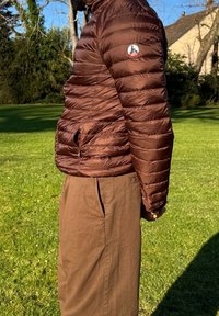 Braune Pufferjacke mit gesteppter Optik, hohem Kragen und Seitentaschen, getragen über braunen Hosen in einer Außenszene.