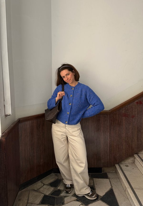 Kobieta stoi na kafelkowej podłodze przy schodach, ubrana w niebieski sweter zapinany na guziki, kremowe szerokie spodnie, sneakersy i trzyma brązową torbę na ramieniu.