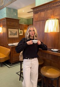 Joven mujer con suéter negro y pantalones blancos bebe de una taza en un bar de café de madera con taburetes y luz cálida.