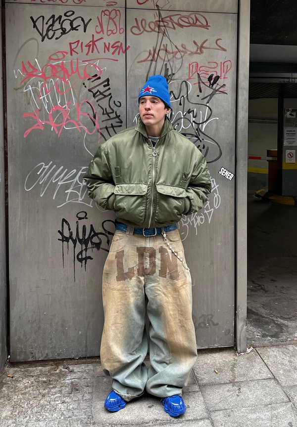 Jaunas vyras mėlynu megztiniu šalmuku, per didelę žalią striukę ir platėjančiomis kelnėmis su užrašu „LDN“ stovi prie sienos, padengtos grafičiais.