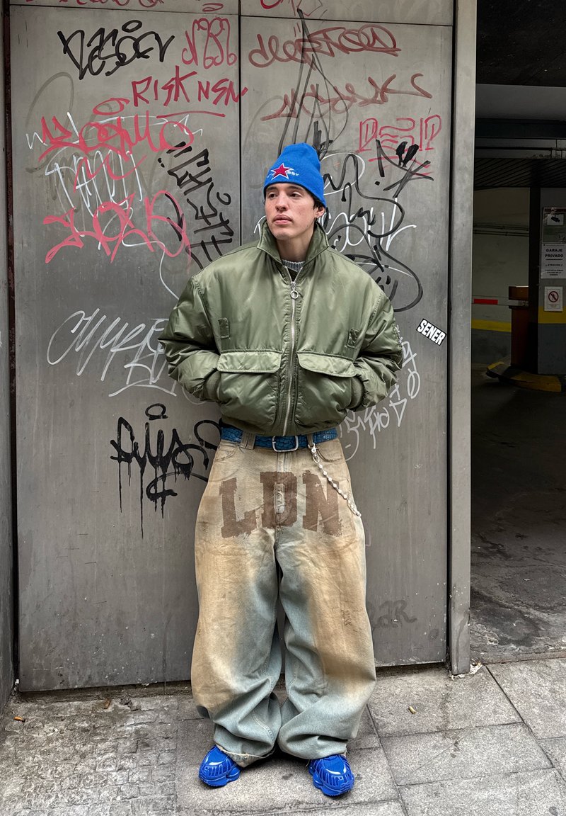 Mladić u plavoj kapi, prevelikoj zelenoj jakni i širokim hlačama s natpisom "LDN" stoji naslonjen na zid prekriven grafitima.