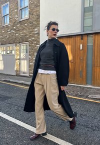 Svart ullfrakk over en grå høyhalset genser, beige plisserte bukser og burgunder hælsko. Tilbehør inkluderer smale solbriller og et belte.