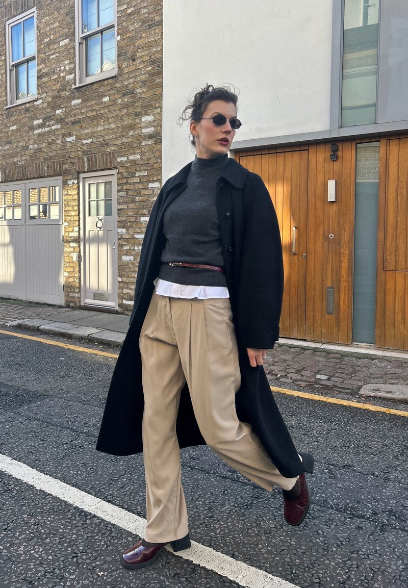 Casaco preto de lã sobre uma camisola de gola alta cinzenta, calças plissadas bege e sapatos de salto bordeaux. Os acessórios incluem óculos de sol estreitos e um cinto.
