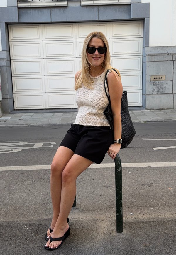 Femme portant des lunettes de soleil, un haut en maille sans manches et un short noir, assise sur un poteau en métal au bord de la rue avec un grand sac noir sur l'épaule.