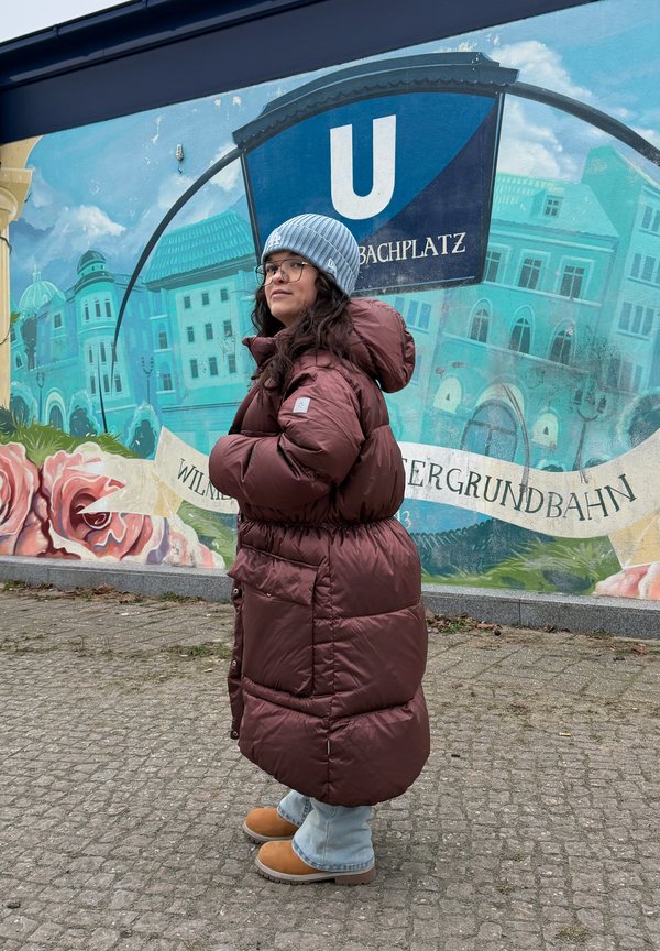 Osoba v hnedej prešívanej bunde, modrej čiapke a topánkach stojí pri nástennom maľbe s nápisom "U" pri stanici metra a mestským pozadím.