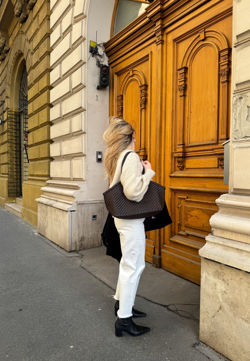 Žena s dlouhými blond vlasy drží tmavý pletený koš, stojí u velkých zdobených dřevěných dveří zasazených do kamenné budovy na chodníku ve městě.