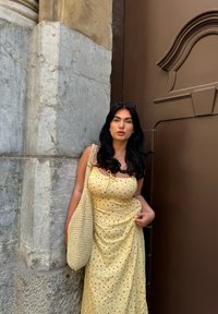 Κίτρινο φόρεμα με λουλουδάτο μοτίβο και στρογγυλή λαιμόκοψη, διαθέτει ρυθμιζόμενους κόμπους. Συνδυασμένο με ένα υφαντό μπεζ τσάντα μπροστά από μια καφέ πόρτα.