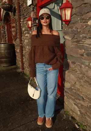 Mujer con suéter marrón off-the-shoulder, jeans azul claro, zapatos beige de plataforma, bolso crema, gafas de sol y gorra blanca, de pie junto a un muro de piedra y una puerta roja.