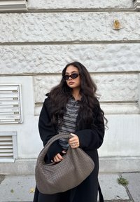 Saco hobo de couro com textura trançada em cinza. O modelo usa um casaco preto sobre uma blusa riscada. O fundo apresenta uma parede branca texturizada.