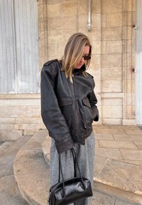 Schwarze Bomberjacke aus Leder mit sichtbaren Nähten, vorderen Taschen und hohem Kragen. Kombiniert mit grauen Hosen im Hahnentrittmuster und einer schwarzen Handtasche.