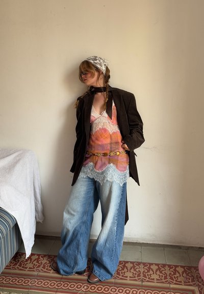 Blazer de pana marrón sobre una blusa de encaje multicolor con bordes en picos, combinada con unos jeans de mezclilla azul claro sueltos y botas marrones.