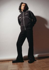Μαύρο bomber μπουφάν με λευκές λεπτομέρειες, φερμουάρ μπροστά και πλευρικές τσέπες, συνδυασμένο με μαύρα φαρδιά παντελόνια και χοντρές μαύρες μπότες.