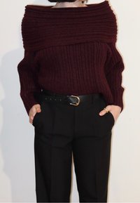 Femme portant un pull bordeaux côtelé à épaules dénudées, rentré dans un pantalon noir taille haute avec une ceinture noire, les mains dans les poches.