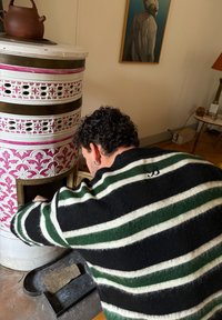 Poêle avec un design floral blanc et rose, forme ronde, et trous apparents ; personne en pull rayé noir et vert ajoutant du bois.