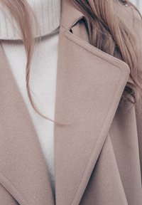 Femme portant un manteau en laine beige sur un pull à col roulé en tricot blanc, avec de longs cheveux châtain clair visibles autour du col.