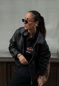 Μαύρο δερμάτινο bomber jacket, μαύρη μπλούζα t-shirt με λογότυπο, χρυσά σκουλαρίκια κρίκοι, μαύρα γυαλιά ηλίου, μαύρα ακουστικά και καφέ τσάντα ώμου.