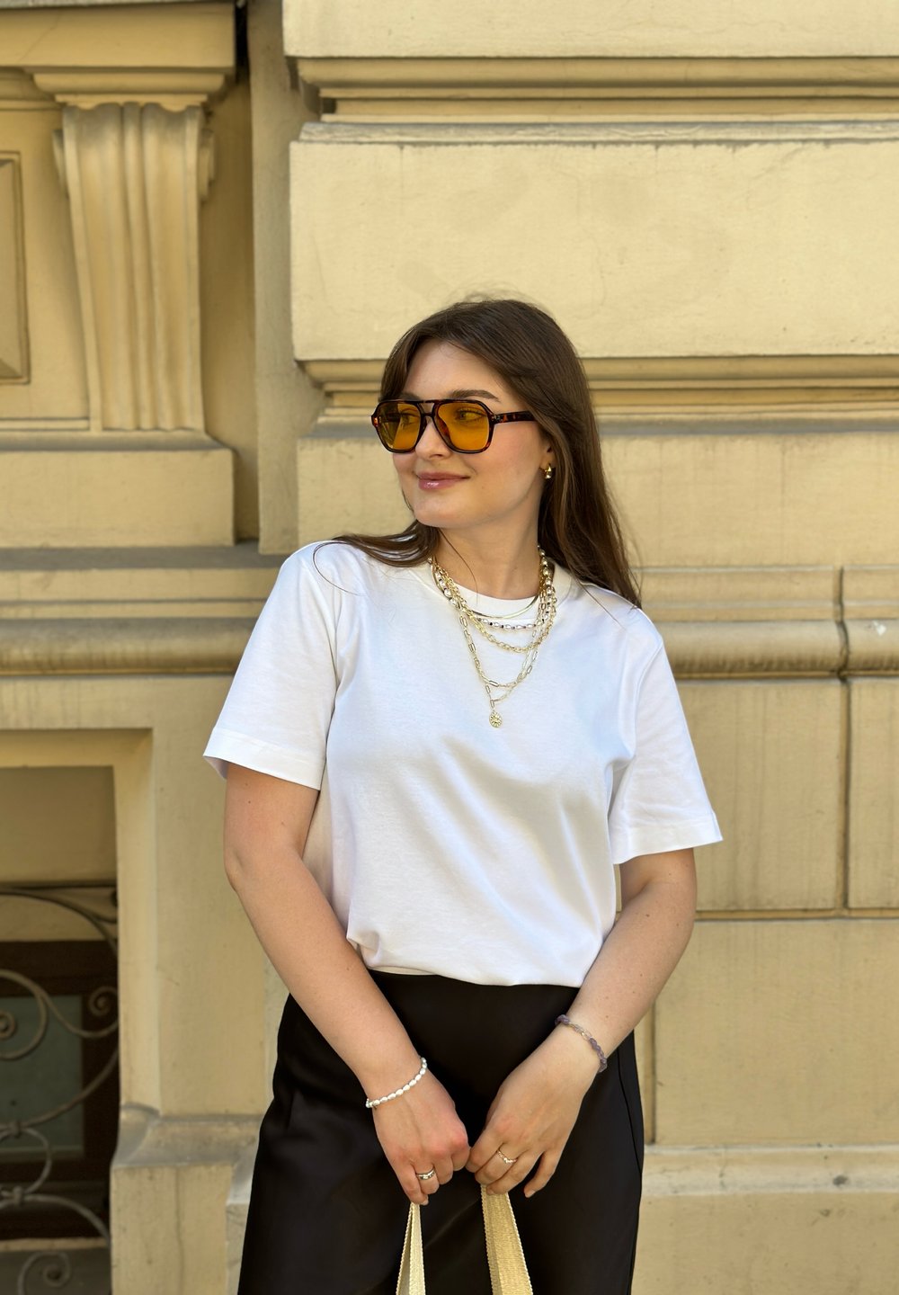 Weißes T-Shirt, schwarze Hose, geschichtete goldene Ketten und große braun-gelbe Sonnenbrille. Hält eine beige Tasche, die Haare sind lang und braun.