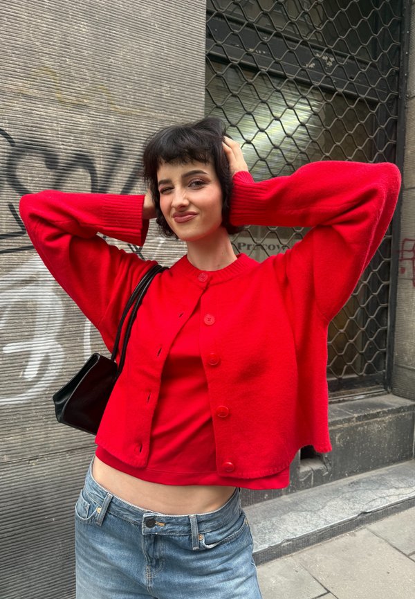 Jeune personne en pull rouge et top pose avec les mains derrière la tête, portant un jean bleu et un petit sac à épaule noir, debout près d’un mur de graffiti.