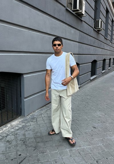 Camiseta blanca, pantalones de pierna ancha color crema y chanclas negras. Bolsa de mano a rayas colgada del hombro. Fondo urbano con paredes grises.