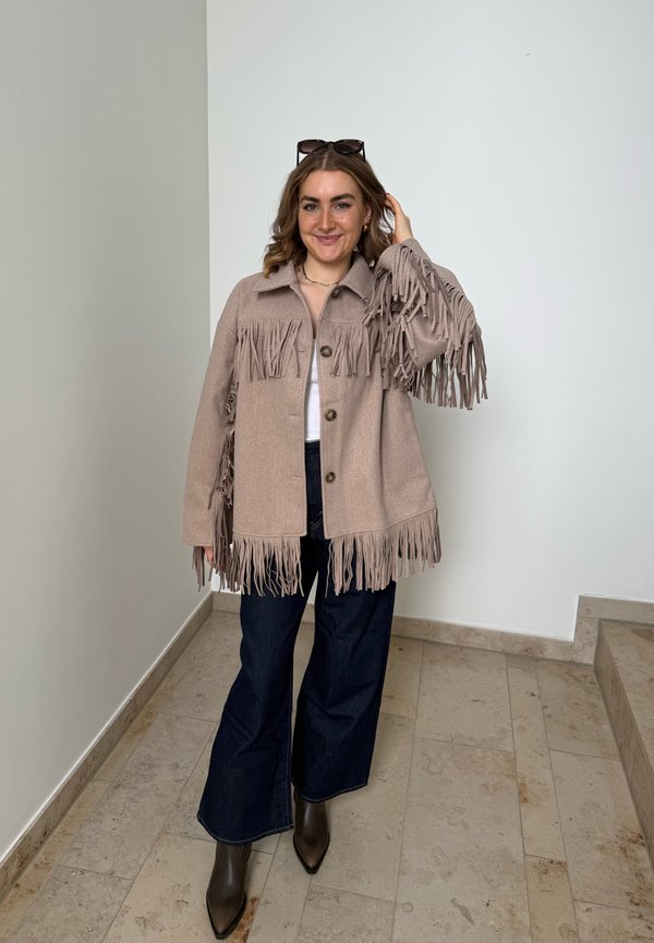Femme debout à l’intérieur portant une veste beige à franges, un pantalon large foncé, des bottes marron, des lunettes de soleil sur la tête, et souriant en touchant ses cheveux.