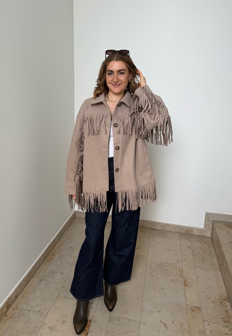 Donna in piedi al chiuso con giacca beige con frange, pantaloni scuri ampi, stivali marroni, occhiali da sole sulla testa, sorridente mentre si tocca i capelli.