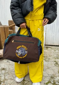 Καφέ και πράσινη τσάντα duffel με κεντητό σχέδιο λιονταριού, χρυσό φερμουάρ και μαύρες λαβέ�ς. Φορεμένη με κίτρινο φόρμα και λευκά αθλητικά παπούτσια.