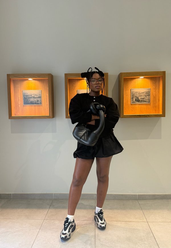 Femme aux cheveux tressés et portant des lunettes, debout bras croisés tenant un sac noir, vêtue d'une tenue noire et de baskets, photos encadrées sur le mur derrière.