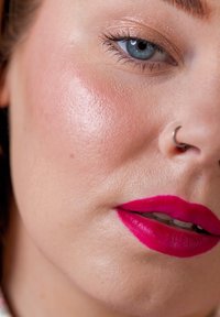 Close-up van een gezicht met een gladde, lichte huid met een matte afwerking, felroze lippenstift, een zilveren neusring en blauwe ogen.