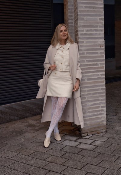 Conjunto de chaqueta y falda de tweed de color claro con un cuello volado, combinado con medias de encaje blancas y mocasines beige, complementado con un abrigo largo.