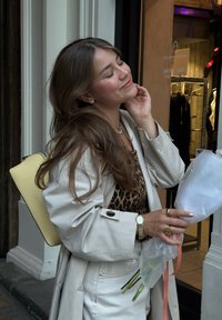 Lys beige trenchcoat over en leopardmønstret topp; holder hvite blomster emballert i klart plast. Gul veske og gulltilbehør synlige.