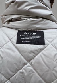 Veste matelassée beige vue de dos avec une étiquette de marque Ecoalf noire indiquant un mode de vie durable et le slogan "Parce qu'il n'y a pas de planète B".