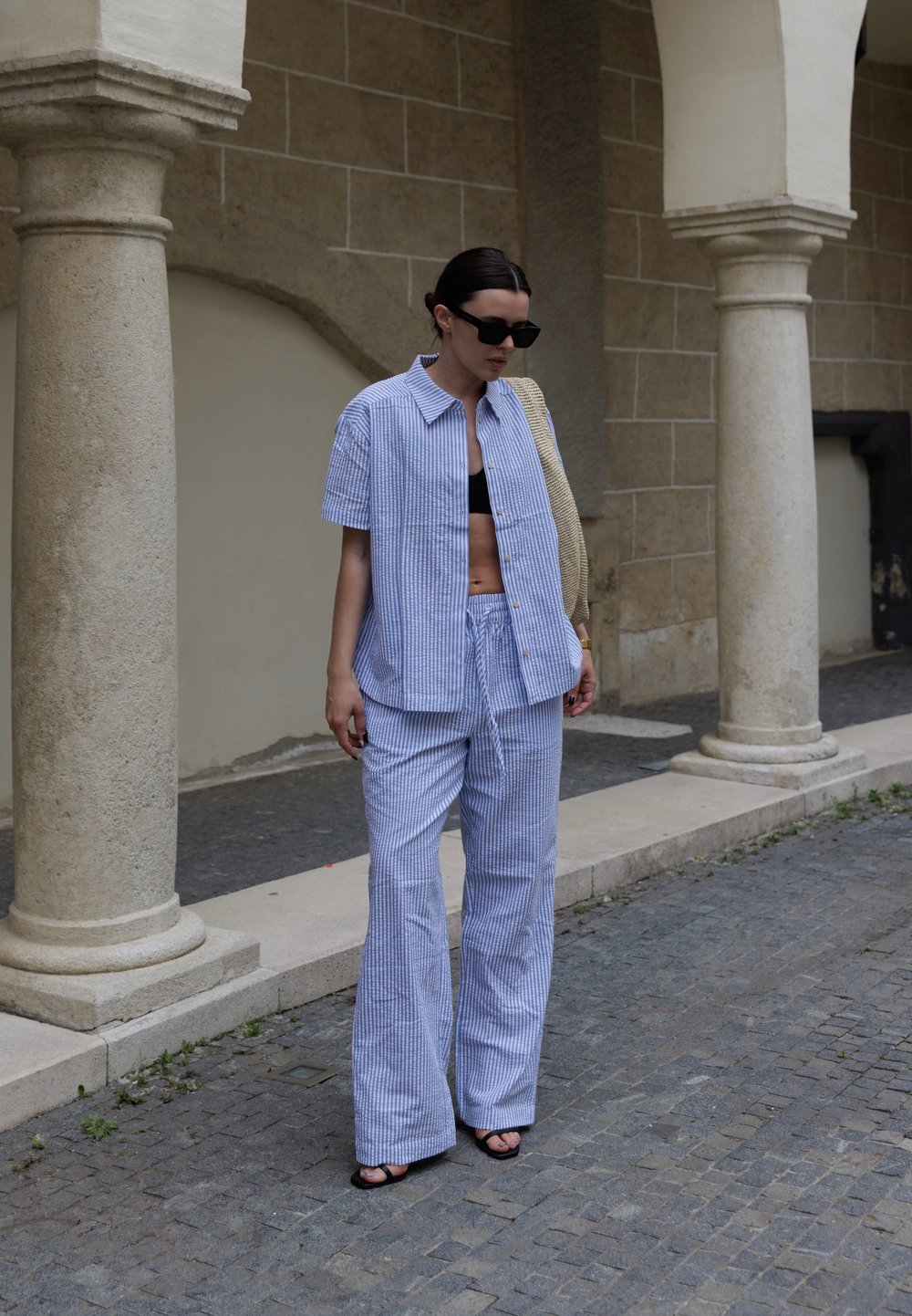 Modra in bela prugasta srajca s kratkimi rokavi ter široke hlače iz lahkega materiala, v kombinaciji s črnimi sandali in sončnimi očali.