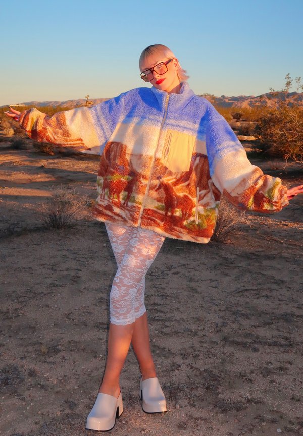 Fleecejakke med et blåt, cremefarvet og brunt landskabsmønster med heste. Kombineret med hvide blonderleggings og chunky hvide plateau sko.