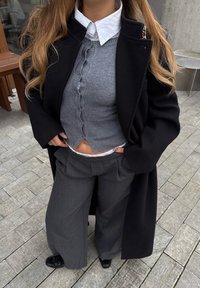 Personne portant un manteau noir par-dessus un cardigan boutonné gris et une chemise blanche, avec un pantalon large gris et des chaussures noires, debout sur un trottoir carrelé.