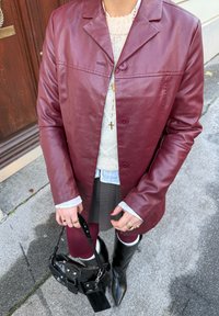Veste en cuir bordeaux avec un col à revers, deux boutons, associée à un pull, une jupe grise et des bottes noires montantes jusqu'aux genoux. Accessoires comprenant un sac à main noir.