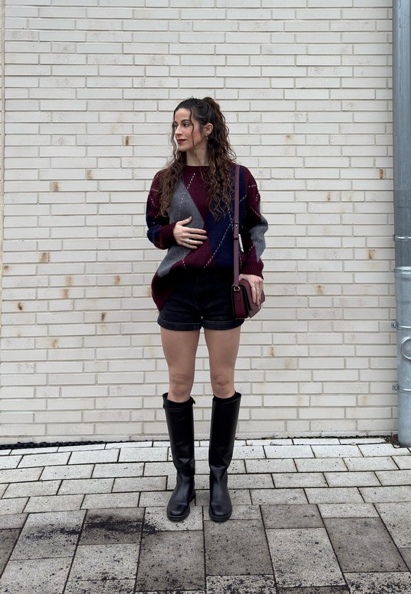 Młoda kobieta z długimi kręconymi włosami, ubrana w wzorzysty sweter, czarne szorty, wysokie kozaki do kolan oraz torebkę przewieszoną przez ramię, stoi przy białej ceglanej ścianie.