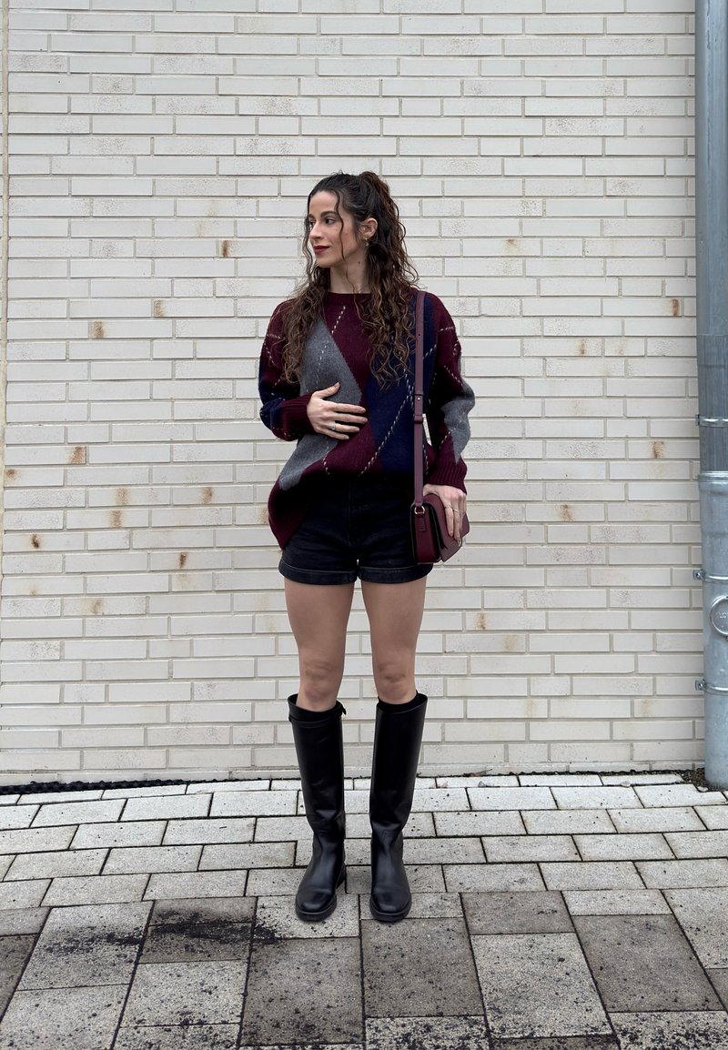 Nuori nainen, jolla on pitkät kiharat hiukset, kuvioitu pusero, mustat shortsit, polven korkeudelle ulottuvat saappaat ja olkalaukku, seisomassa valkoista tiiliseinää vasten.