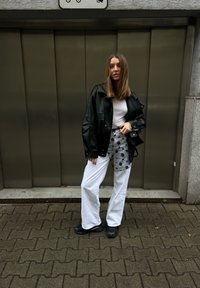 Γυναίκα που στέκεται σε πεζοδρόμιο μπροστά από μια μεταλλική πόρτα, φορώντας μαύρο δερμάτινο μπουφάν, λευκό πουκάμισο, λευκό παντελόνι με μαύρο λουλουδάτο σχέδιο και μαύρα παπούτσια.