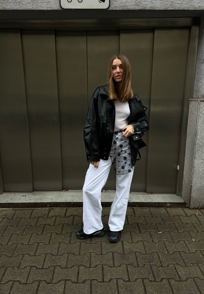 Mujer de pie en una acera frente a una puerta metálica, vestida con una chaqueta de cuero negra, camisa blanca, pantalones blancos con parche floral negro y zapatos negros.
