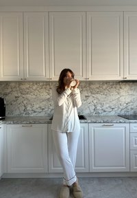 Camicia bianca a maniche lunghe e pantaloni con piping nero, in piedi in una cucina moderna con backsplash in marmo e armadietti chiari, che tiene una tazza.