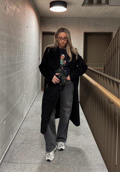 Abrigo largo negro sobre una sudadera gráfica, combinado con jeans oscuros de pierna ancha y zapatillas plateadas. El fondo presenta un pasillo de concreto y una barandilla.