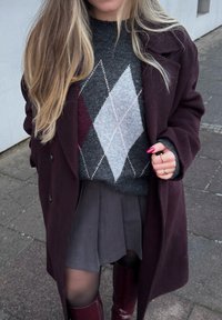 Femme portant un manteau bordeaux par-dessus un pull gris à motif argyle, une jupe plissée grise, des collants noirs, des bottes bordeaux et des bagues en or, debout sur le trottoir.
