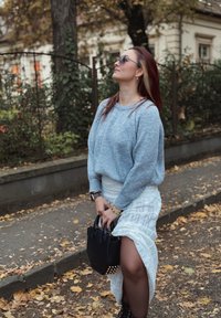 Chunky lichtblauwe gebreide trui, witte textuurs rok met een split en een kleine zwarte handtas met gouden accenten. Herfstsfeer met gevallen bladeren.