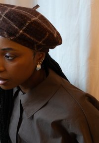 Ruskea ruudullinen villaberet, jossa on pyöreä muoto ja hienovarainen tekstuuri. Korvakorut, joissa on hopeinen ympyräsuunnittelu ja tähden korostus. Ruskea kauluspaita.